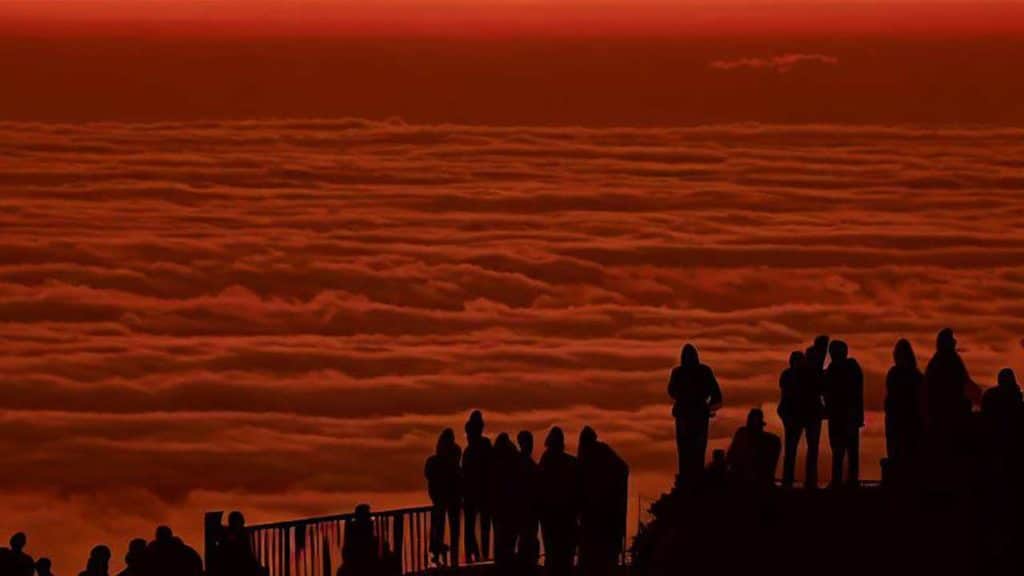 Siluetas de personas de espaldas viendo un atarceder desde un mirador sobre un mar de nubes