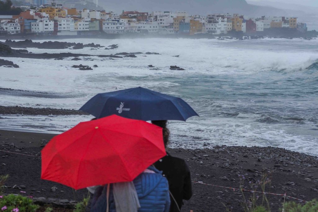 Personas con paraguas junto a la costa del norte de Tenerife, con fuerte oleaje y cielo cubierto
